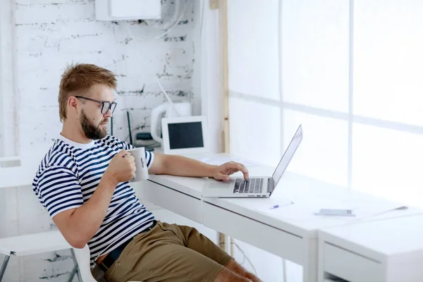 年轻英俊的生意人在办公桌上工作的笔记本电脑坐在办公室喝咖啡 — 图库照片
