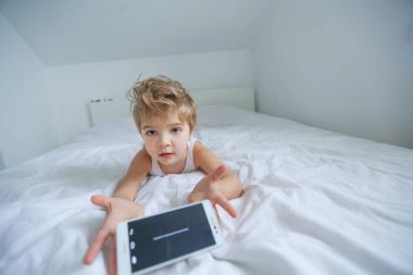 küçük çocuk beyaz yatakta smartphone üzerinde oynarken yalan