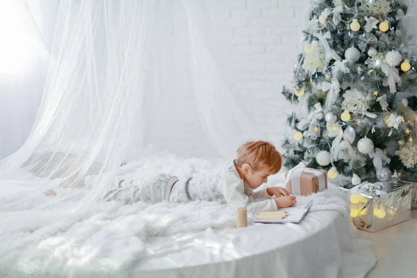 Повнометражний Маленький Хлопчик Лежить Білому Ліжку Біля Ялинки Пише Блокноті — стокове фото