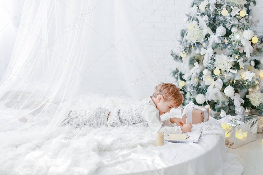 full length of little boy lying on white bed near Christmas tree and writing in notebook