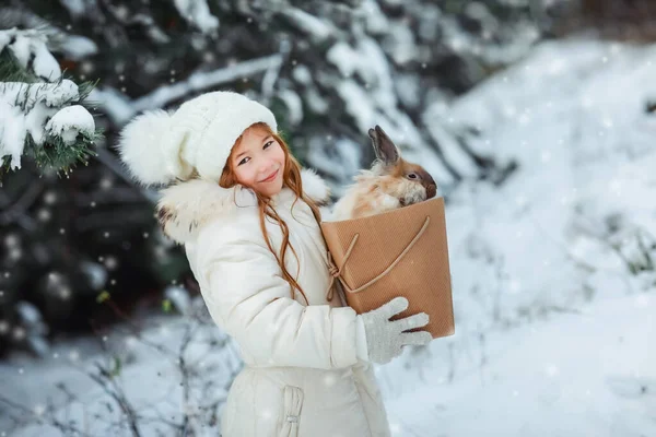 Una chica con una chaqueta blanca y esponjosa en un bosque cubierto de nieve sostiene un conejo vivo decorativo en una caja — Foto de Stock
