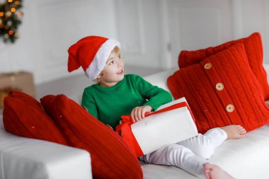 Santaclaus şapkalı ve yeşil kazaklı bir çocuk kanepede oturuyor ve elinde hediyesiyle beyaz bir kutu tutuyor.