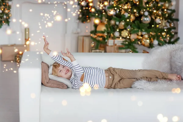 Un băiat cu păr blond pe fundalul unui pom de Crăciun se află pe o canapea și visează la cel mai bun cadou al lui Moș Crăciun — Fotografie, imagine de stoc