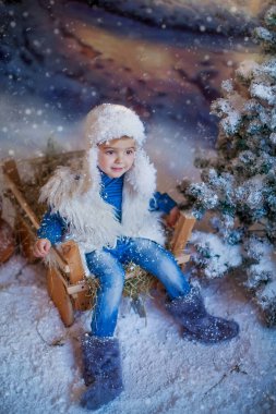 Beyaz şapkalı ve botlu bir çocuk kış köyündeki yapay bir ormanın arka planında.