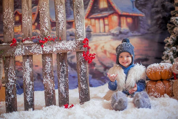 一个小男孩坐在靠近木制篱笆的人造雪地上，屋后有闪闪发光的圣诞装饰品 — 图库照片