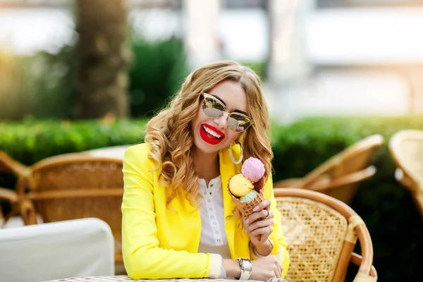 漂亮的金发女人在蛋筒里吃冰淇淋. 戴眼镜的女孩穿着一件黄色的亮夹克. — 图库照片