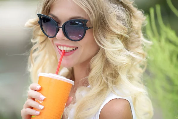 A blonde woman in sunglasses denim pants and a white shirt drinks a cocktail on a hot summer day