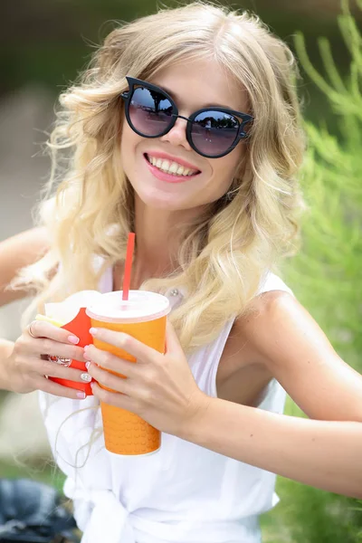 Une femme blonde en pantalon denim lunettes de soleil et une chemise blanche boit un cocktail lors d'une chaude journée d'été — Photo
