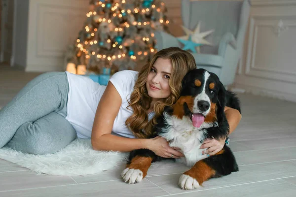 En vacker kvinna med långt hår ligger på en matta mot bakgrund av en julgran och bredvid henne ligger hennes stora hund — Stockfoto