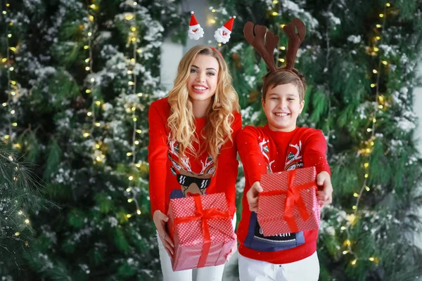 一个留着漂亮头发和红色毛衣的女人站在有一个成年儿子的天然圣诞树旁，手里拿着装有礼物的盒子 — 图库照片