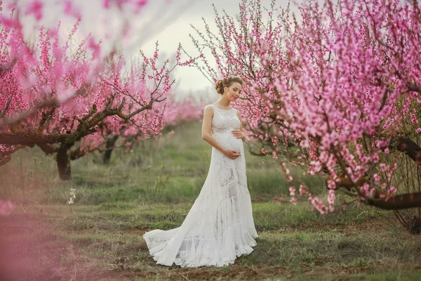 Uma mulher em um vestido de noiva branco longo entre as árvores florescentes de pêssego — Fotografia de Stock