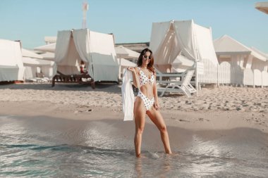 Uzun bacaklı ve seksi formalı bikinili güzel bir kadın Sea Edge 'de rahatlıyor. Şık beyaz mayo giymiş, güneş gözlüğü takan, sağlıklı tenli bir kadın.