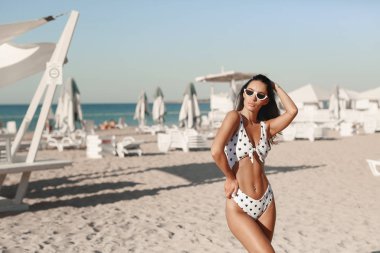 Beyaz bikinili ve bej renkli bir kadın model gün batımında kumlu bir plajda oturuyor. Rahatla, tatil ve yaz tatili.