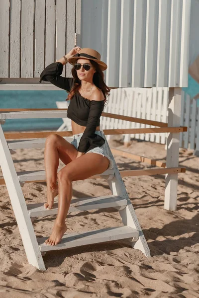 Una mujer se sienta en la arena en la playa cerca de una cerca de madera blanca con gafas de sol y pantalones cortos — Foto de Stock