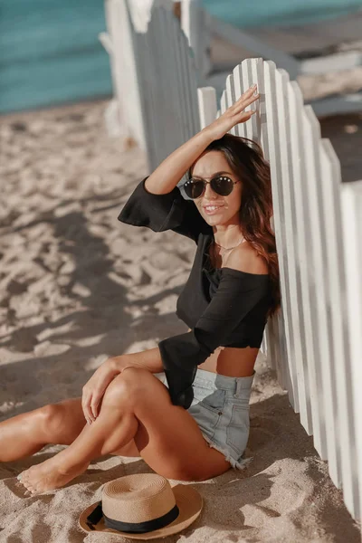一个女人穿着太阳镜和短裤，坐在沙滩上一个白色木制栅栏旁边的沙滩上 — 图库照片