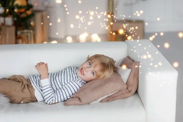 Un niño con el pelo rubio sobre el fondo de un árbol de Navidad yace en un sofá y sueña con el mejor regalo de Papá Noel — Foto de Stock