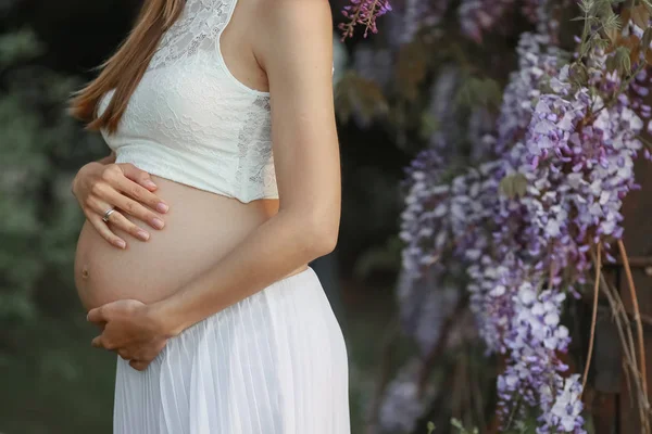 Una donna incinta in gravidanza tardiva nel giardino di glicine in fiore abbraccia la pancia — Foto Stock