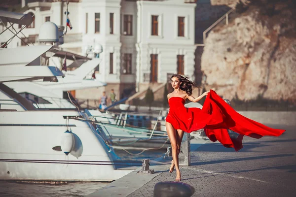 Modelo menina em um longo vestido vermelho fluindo caminha ao longo da orla com iates — Fotografia de Stock