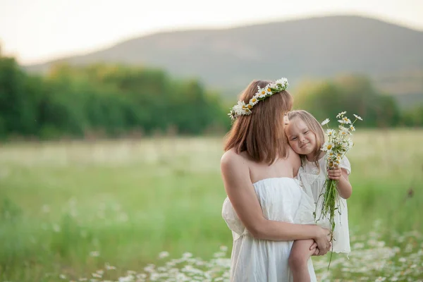 Foto de cerca de una madre con un vestido blanco y un bebé en sus brazos en un campo con hierba verde en el fondo de las montañas —  Fotos de Stock