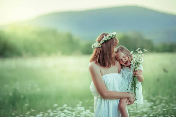 Foto de cerca de una madre con un vestido blanco y un bebé en sus brazos en un campo con hierba verde en el fondo de las montañas —  Fotos de Stock