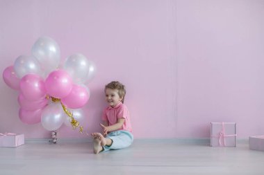 Doğum günü kutlamasında çıplak ayaklı bir çocuk pembe ve beyaz balonlarla çevrili oturur.