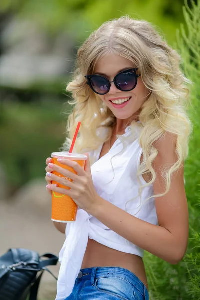 A blonde woman in sunglasses denim pants and a white shirt drinks a cocktail on a hot summer day