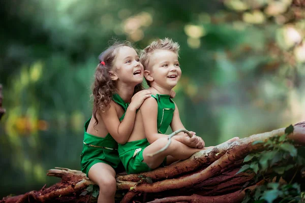 Niños un niño y una niña en el bosque cerca de un fabuloso lago verde sentado en un árbol caído —  Fotos de Stock