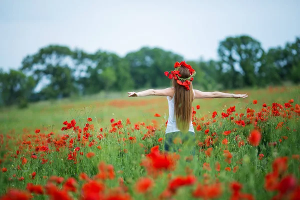 Una mujer con una camisa blanca y pantalones de mezclilla con una corona de flores rojas en la cabeza en un campo —  Fotos de Stock