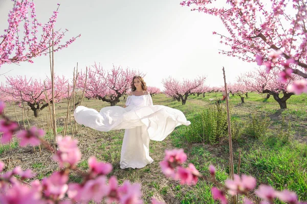 Güzel beyaz elbiseli yetişkin bir kadın pembe çiçekli ağaçlarla dolu bir bahçede koşuyor. — Stok fotoğraf