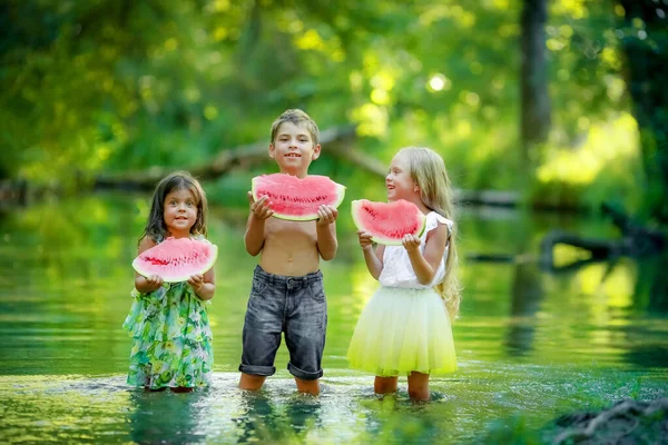 Tre bambini all'aperto stanno insieme nel lago e tengono in mano una gomma da cancellare di un succoso anguria — Foto Stock