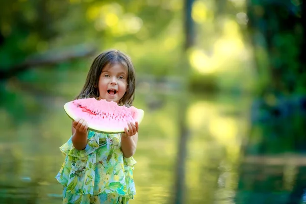 Une fille d'âge préscolaire se tient près sur le fond de la rivière avec un fond flou et mange une pastèque juteuse — Photo