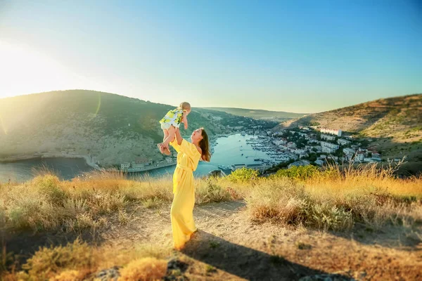 Egy hosszú sárga ruhába öltözött anya áll egy babával a karjában a hegy tetején. — Stock Fotó