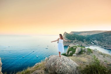 Mavi elbiseli bir kadın gülümsüyor ve suyun üstündeki bir dağa karşı bir taşın üzerinde duruyor.