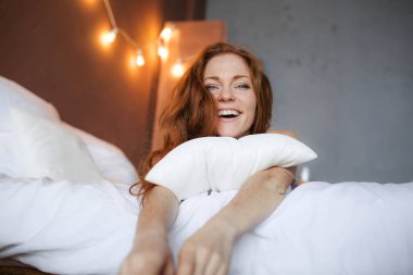 Yatakta sabah, çillileri yatakta yatan, yastığı kucaklayan, gülümseyen, sabahın tadını çıkaran, çilli genç, kızıl saçlı genç bir kadın.