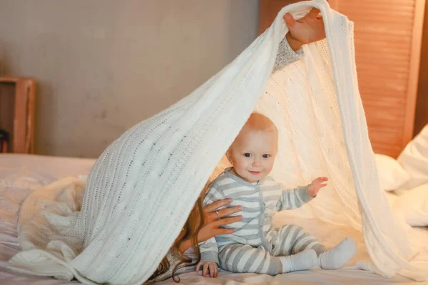 Baby and mom play on the white bed and hide under the covers — Stock Photo, Image