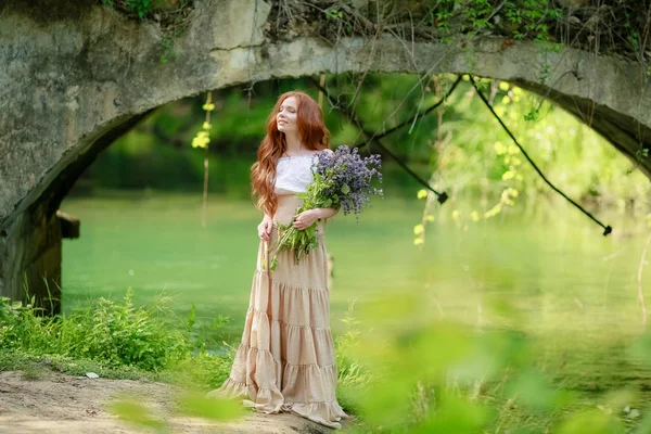 A woman in a long dress walks near the forest old bridge over the river — 스톡 사진