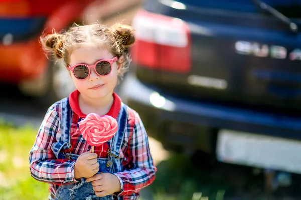 Дівчина в сонцезахисних окулярах і цукерках на паличці в руках на тлі автомобілів — стокове фото