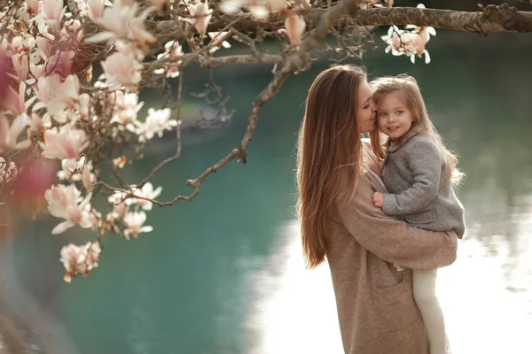 A woman holds a child in her arms against the background of a lake in a garden with magnolia — Stockfoto