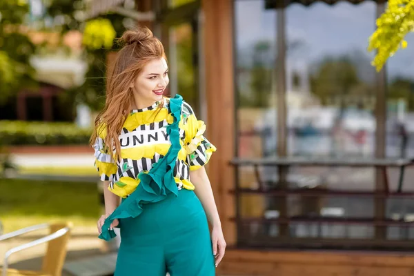 Mujer de cerca se levanta contra el fondo de la cafetería de la ciudad en una camiseta sin mangas de color amarillo brillante y monos de color turquesa — Foto de Stock