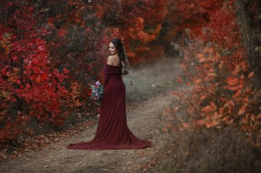 A woman in an elegant red dress and a bouquet of flowers in her hands stands with her back to the camera and looks over her shoulder towards the camera