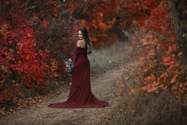 Μια γυναίκα με ένα κομψό κόκκινο φόρεμα και ένα μπουκέτο λουλούδια στα χέρια της στέκεται με την πλάτη της στην κάμερα και κοιτάζει πίσω από τον ώμο της προς την κάμερα — Φωτογραφία Αρχείου