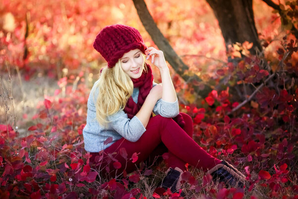 A woman in a warm knitted hat sits under a tree with fallen bright red natural leaves.