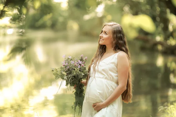 長い白いドレスの妊婦は川の近くの森の中を裸足で歩く — ストック写真