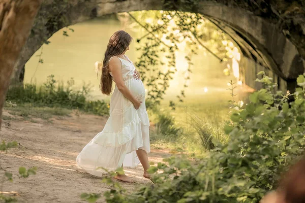 Těhotná žena v dlouhých bílých šatech chodí bosá v lese v blízkosti řeky — Stock fotografie