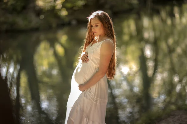 Těhotná žena v dlouhých bílých šatech chodí bosá v lese v blízkosti řeky — Stock fotografie
