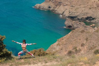 Spor kıyafetleri giymiş bir kadın denizin üstündeki dağın tepesinde egzersiz yapıyor.