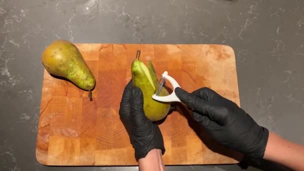 Vrouw in zwarte keuken handschoenen reinigt peer schil met mes snijden — Stockvideo