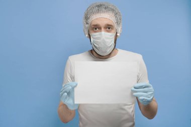 Beyaz tişörtlü, tek kullanımlık şapkalı, tıbbi maskeli ve eldivenli bir adam mavi arka planda beyaz bir kağıt tutuyor.