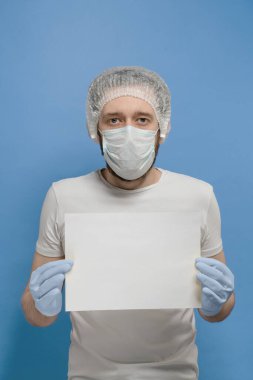 Beyaz tişörtlü, tek kullanımlık şapkalı, tıbbi maskeli ve eldivenli bir adam mavi arka planda beyaz bir kağıt tutuyor.