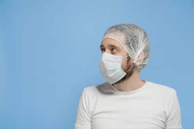Beyaz tişörtlü ve tıbbi maskeli bir adam mavi bir arka plana bakıyor.